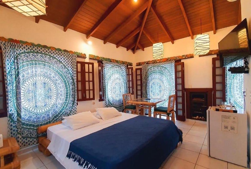 Quarto da Pousada Pura Vida Maresias com uma cama de casal no meio. Em frente a cama está um frigobar e ao lado dela está uma mesa de dois lugares. Uma tv está também virada para a cama. Existem várias janelas e portas espalhados pelo quarto. Essa também é uma boa opção de pousadas pet friendly em Maresias.