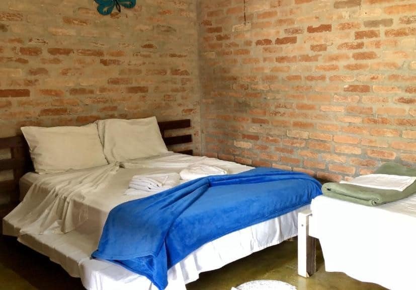 Quarto da Pousada Raio de Sol Maresias. Uma cama de casal está no meio, em frente a uma cama de solteiro. As paredes são de tijolos.