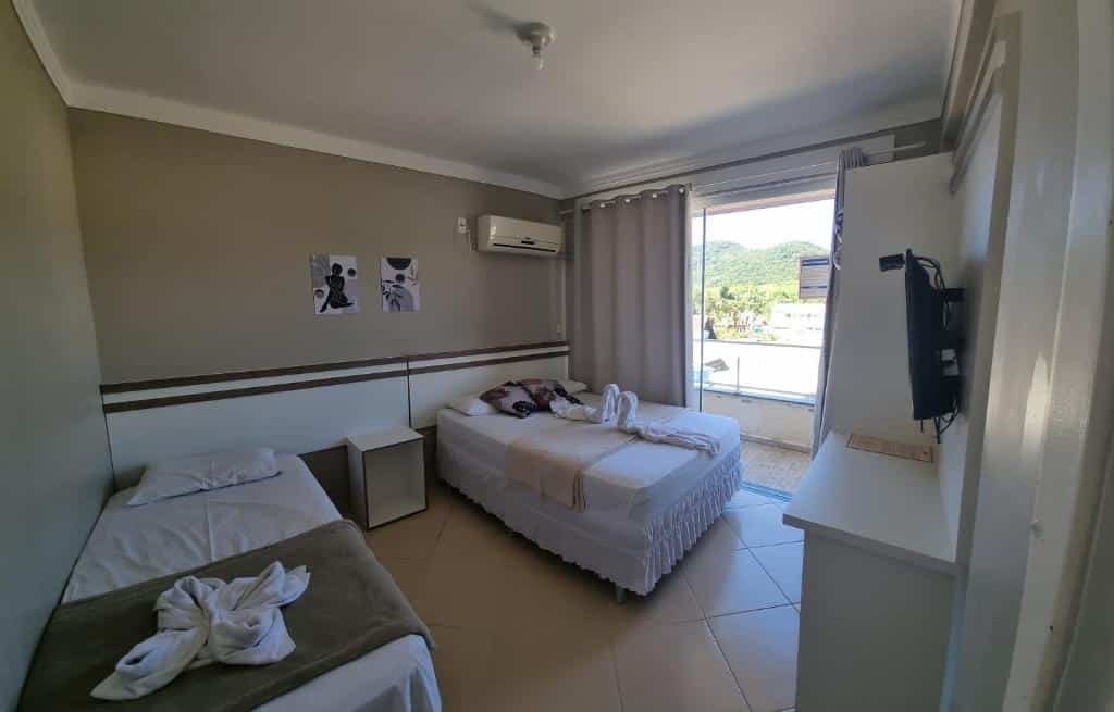 Quarto da Pousada Recanto do Cowboy. Uma cama de solteiro do lado esquerdo, do lado direito uma cama de casal, um ar-condicionado e a varanda do quarto. De frente para as camas uma televisão. Foto para ilustrar post sobre pousadas pet friedly em Santa Catarina.