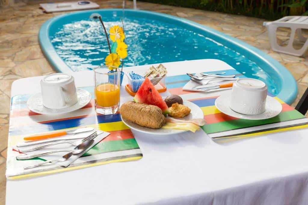Uma mesa posta com café da manhã para duas pessoas, no fundo a piscina da Pousada Recanto Domus.