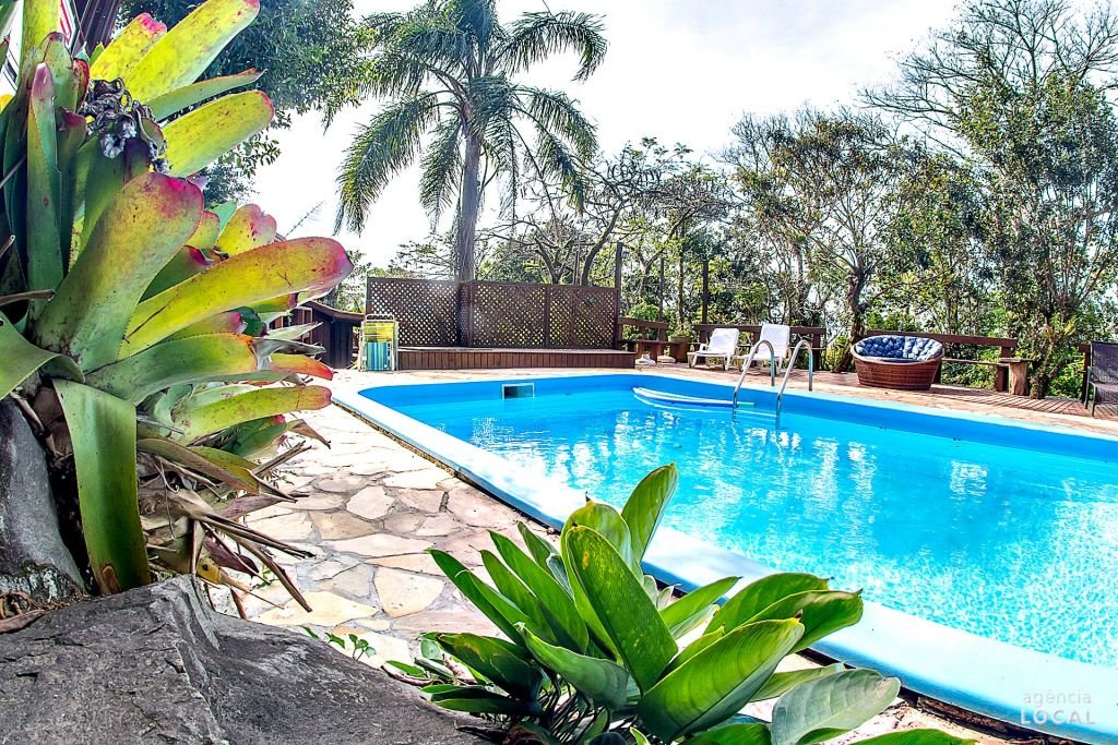 Área externa da Pousada Rosa. Umas plantas do lado esquerdo, do lado direito uma piscina com cadeiras de tomar sol, sofá e árvores.