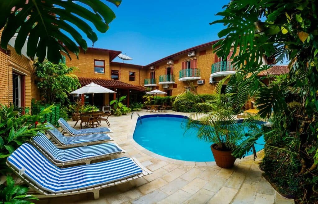 Piscina do Pousada Villa Di Rimini com um deck ao redor cheio de vasos de plantas, espreguiçadeiras e guarda-sóis brancos