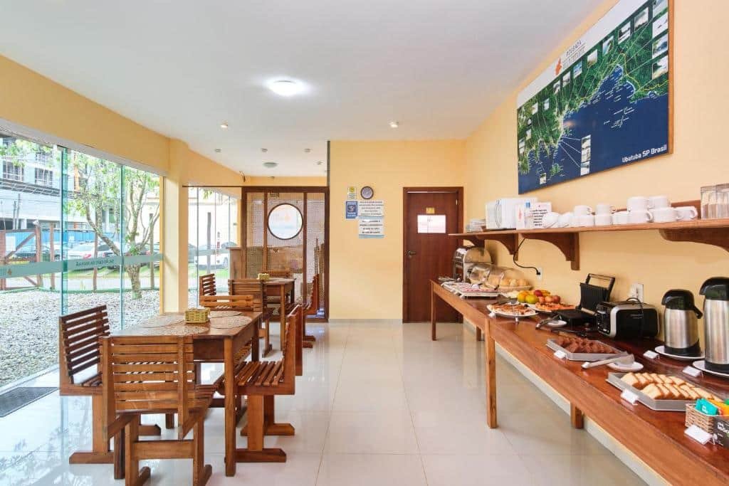 Área de refeição da Pousada Villa Piemonte. Do lado direito uma mesa com pães, bolos, frutas, utensílios de cozinha e garrafas térmicas. No meio cadeiras e mesas de madeira, do lado esquerdo uma porta de vidro.