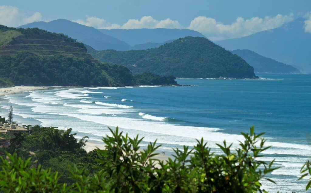 Vista de Juquehy. Tirada entre as árvores, a foto mostra as ondras quebrando na praia e montanhas distantes cheias de vegetação.
