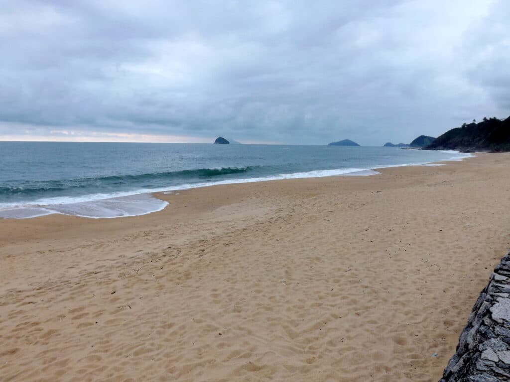 Boiçucanga deserta com ondas suaves, areia dourada e fofa e sob um céu nublado. À distância, pequenas ilhas são visíveis no horizonte.