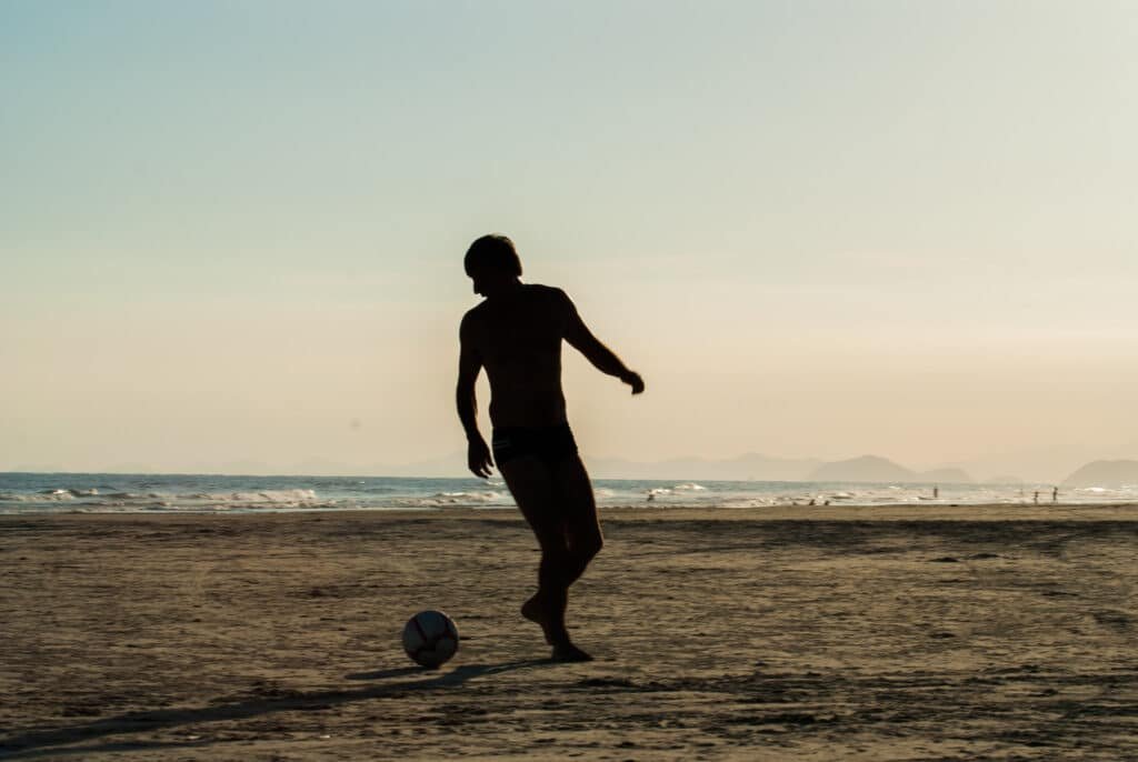 Silhueta de uma pessoa jogando futebol ns Praia da Boraceia ao pôr do sol, com ondas e colinas distantes ao fundo.