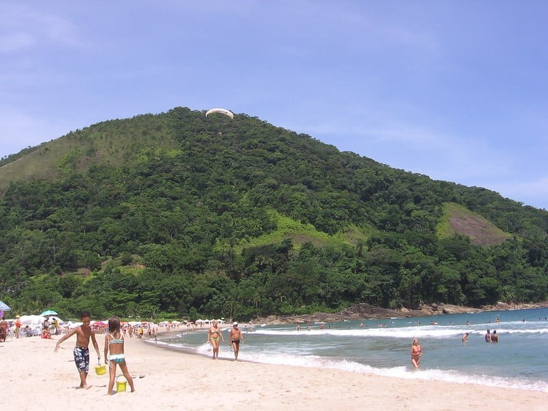 Praia de Camburizinho com pessoas tomando sol e nadando. Uma colina florestada se ergue ao fundo sob um céu azul claro.