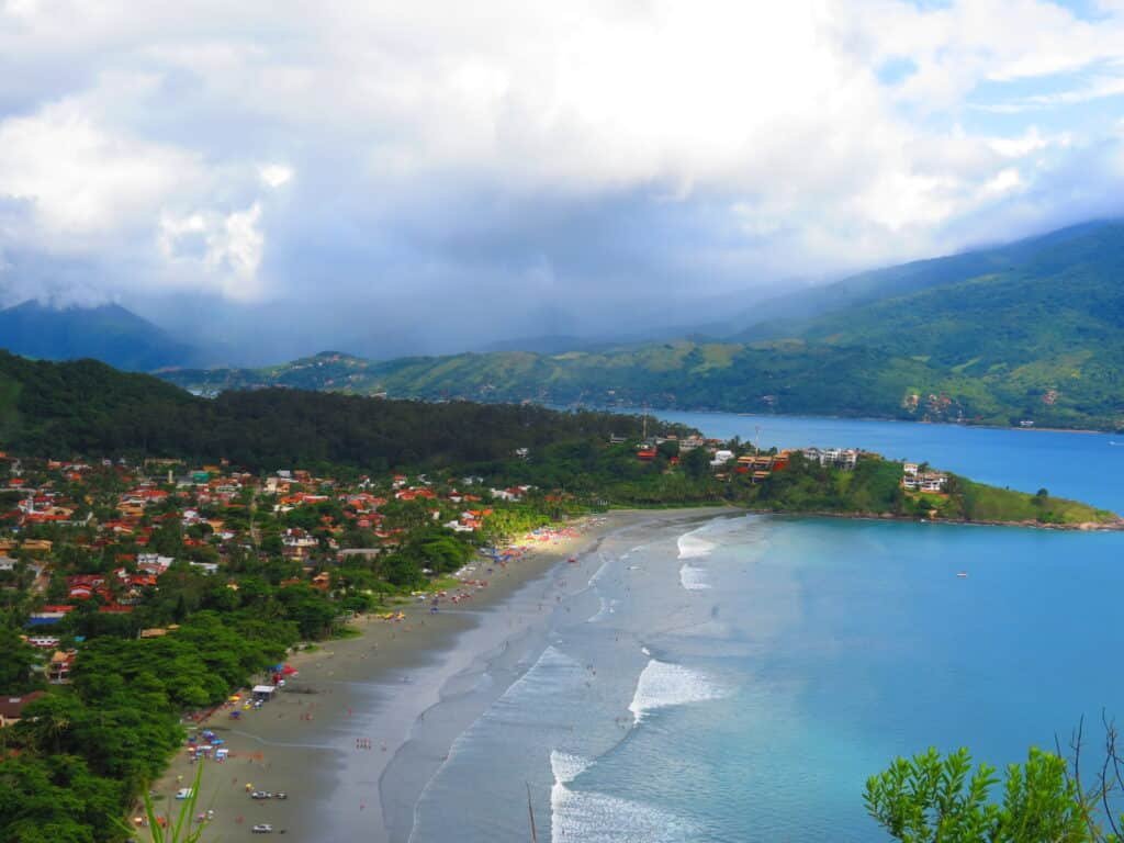 Vista aérea de Barequeçaba. A praia está do lado direito, com areia curva e colinas verdes ao redor, sob um céu parcialmente nublado. Do lado esquerdo está a vegetação e várias casas.