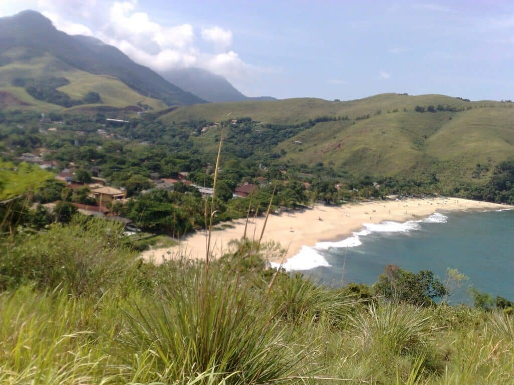 Foto de longe da Praia de Paúba. Ela tem ondas suaves, cercada por morros verdes e casas espalhadas sob um céu limpo.