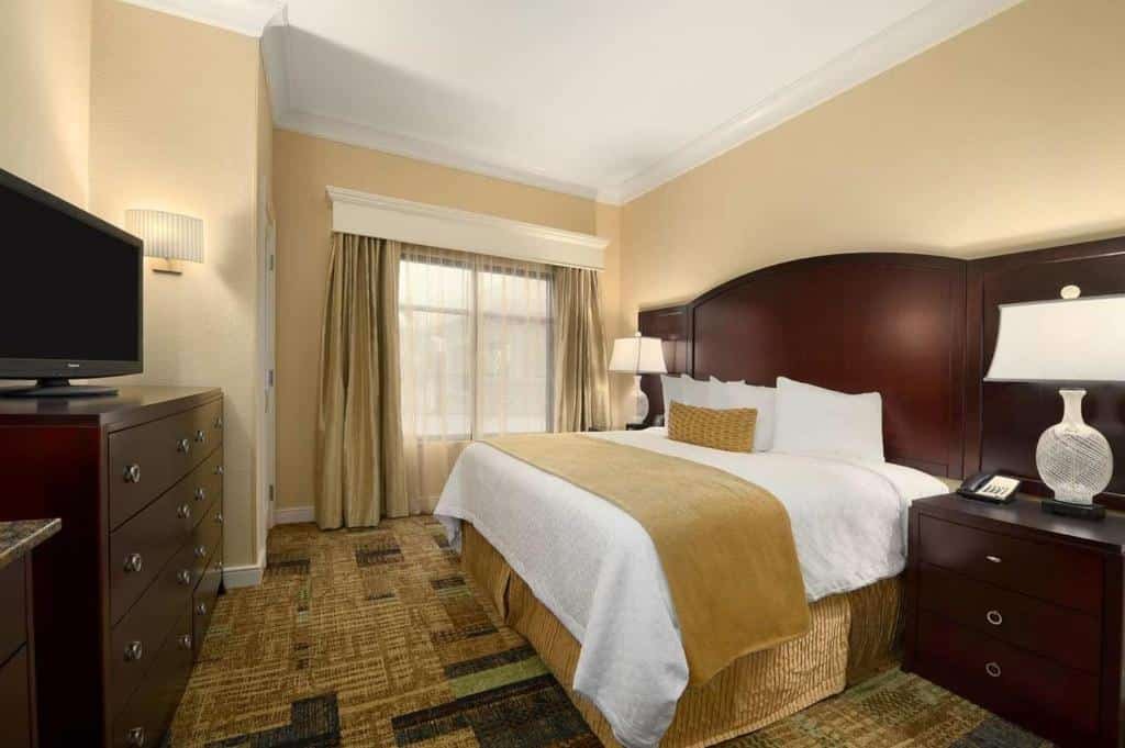 quarto do Embassy Suites by Hilton Orlando Lake Buena Vista South com janelas e cortinas com cômoda e tv em frente à uma cama de casal com mesinha e luminária em ambos os lados com detalhes em madeira