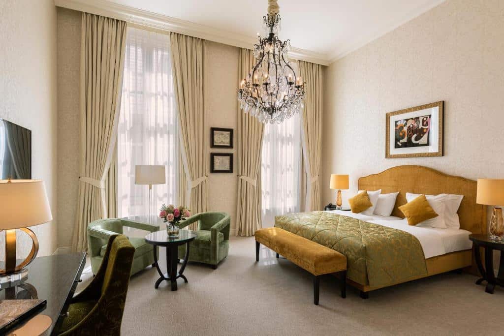 quarto com cama de casal do Grand Hotel Casselbergh Brugge com lustre, duas janelas grandes com cortina, duas poltronas e ainda mesa de trabalho com TV