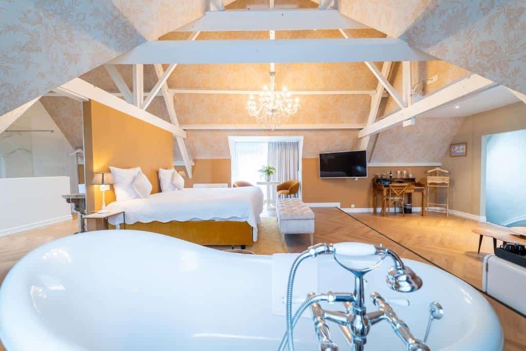 quarto amplo do Grand Hotel Normandy que conta com uma banheira, cama de casal, área de banho atrás da cama, uma tv em frente à banheira, uma mesinha e a porta na frente da cama