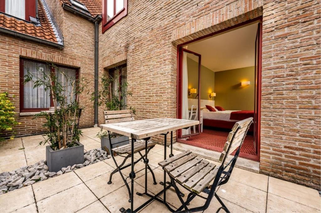 varanda com mesa e cadeiras de madeira com alguma grama do Hotel Acacia com vista para o quarto com cama de casal em detalhes em vermelho, as paredes externas são de tijolinho à vista