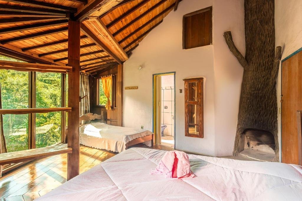 Vista do quarto da Pousada Alquimia com camas, lareira e janelas amplas com vista para árvores.