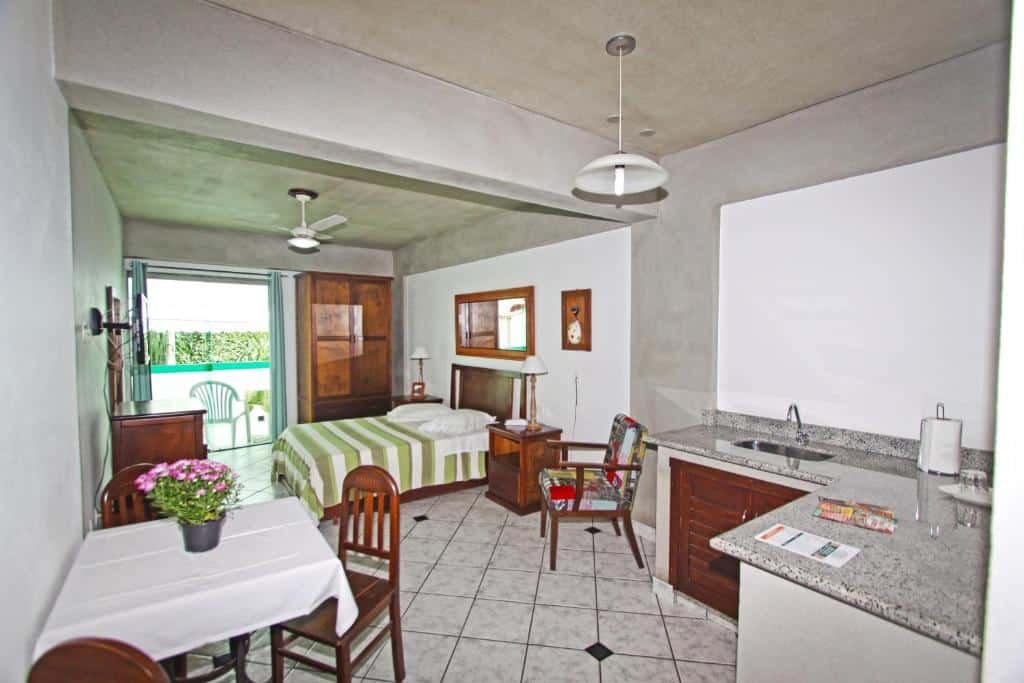 Quarto da hospedagem Aventureiro pousada em Teresópolis com cama de casal, mesa com cadeiras, pia de lavar louça, ventilador de teto e sacada ao fundo, ilustrando post pousadas em Teresópolis.