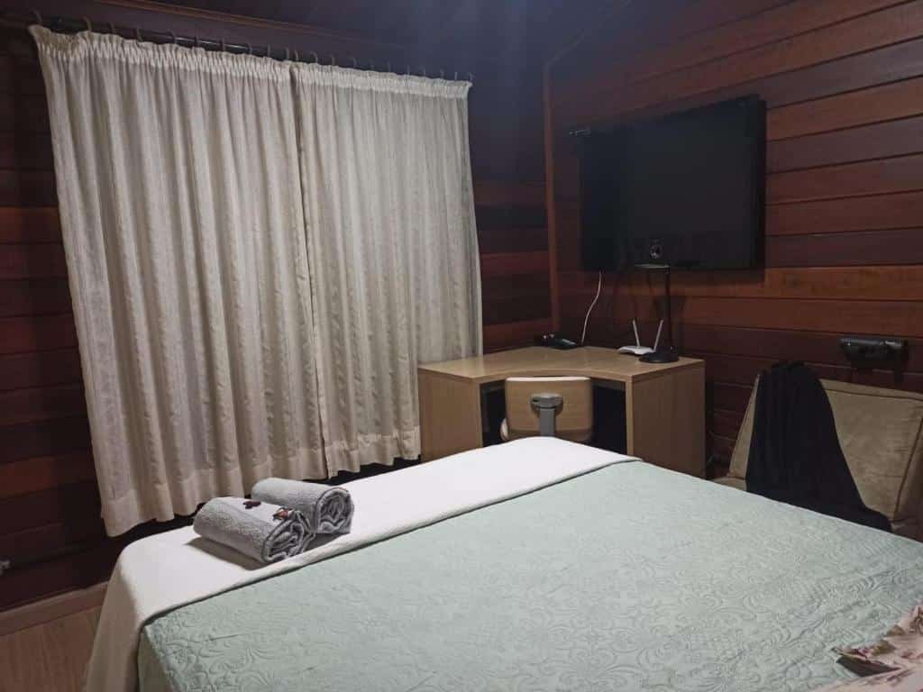 Quarto no Belas Águas Mairiporã. Uma cama de casal de frente pra janela com cortina, uma mesa de trabalho e uma televisão.