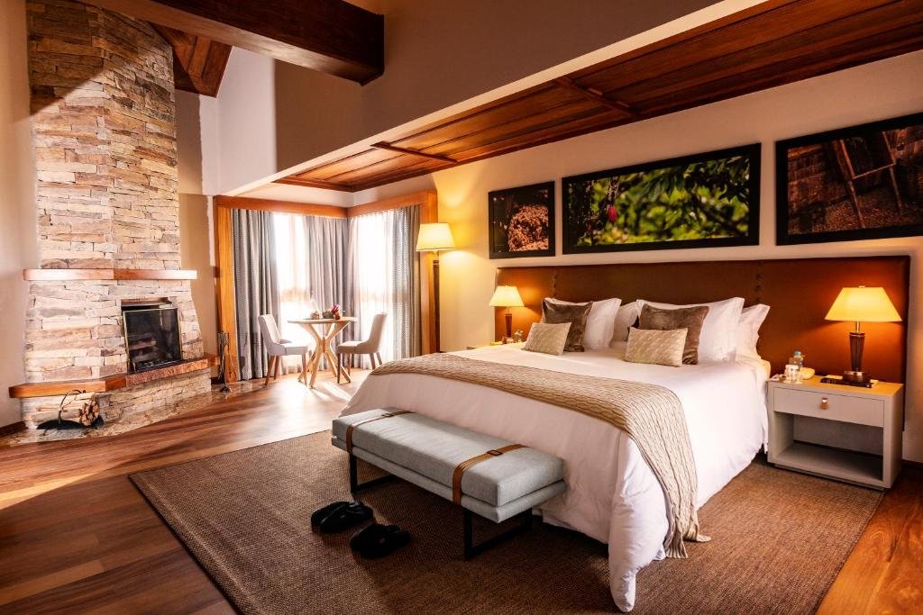 Quarto no Bendito Cacao Resort & Spa com uma lareira de pedra, uma cama de casal, espaço amplo, duas janelas, alguns abajures, chão de madeira com um tapete e uma mesinha com dois lugares