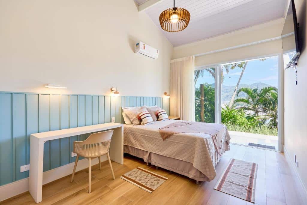 Quarto da Casa de alto padrão com ampla vista para o mar com cama de casal do lado esquerdo da imagem no centro do quarto e uma cômoda do lado esquerdo da cama e do lado direito da cama uma mesa com cadeira.