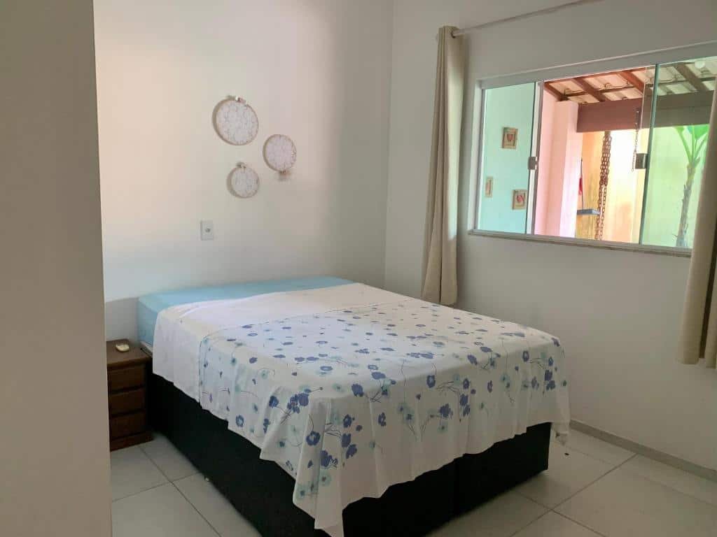 Quarto do airbnb Casa de Férias. No meio do quarto está a cama de casal, no canto esquerdo da cama há uma mesa de cabeceira  bem pequena e no lado direito do quarto há uma janela de vidro aberta com cortinas ao lado. Imagem para ilustrar o post airbnb em Itatiaia.