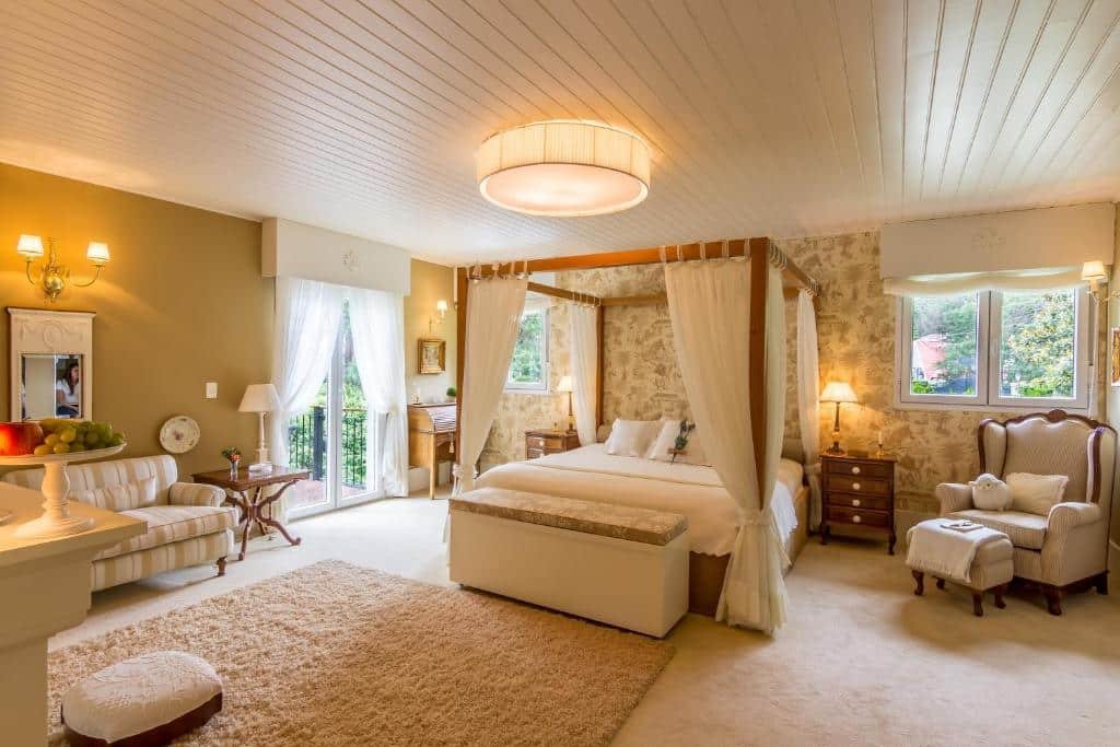 Quarto amplo da Casa Regaleira Hotel Boutique com uma cama de casal com dossel, uma sacada com cortinas brancas, diversas janelas, duas poltronas estofadas, um pequeno sofá, um lustre, entre outros itens de decoração, tudo em tons de bege e madeira