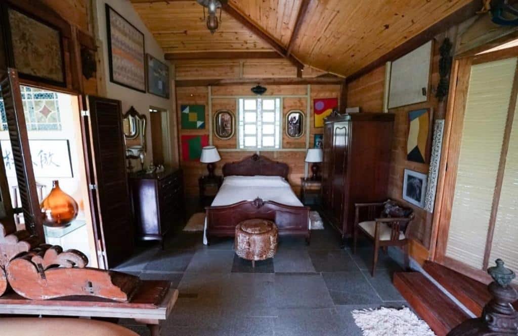 Uma cama de casal no meio, com um puff redondo na frente, no Chalé Portucale. Um armário em cada lado da cama, uma porta do banheiro no lado esquerdo. Bastante luz natural no quarto. Foto para ilustrar post sobre Pousadas em Embu das Artes.