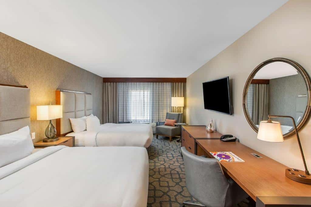 Quarto do DoubleTree by Hilton San Antonio Northwest - La Cantera com uma cama de casal e uma de solteiro, uma janela com cortinas, uma poltrona, um móvel com gavetas, uma televisão na parede de frente com as camas, e uma mesa de escritório com uma cadeira