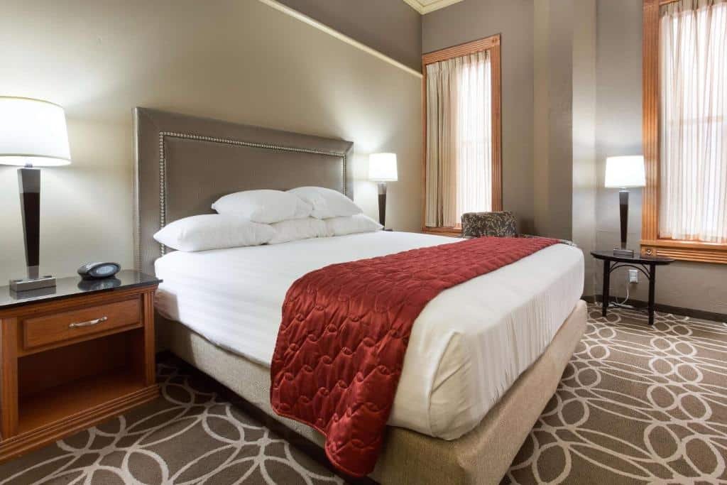Quarto simples do Drury Inn & Suites San Antonio Riverwalk com uma cama de casal, duas janelas com cortinas, duas mesinhas de cabeceira com abajures e carpete marrom com detalhes em branco
