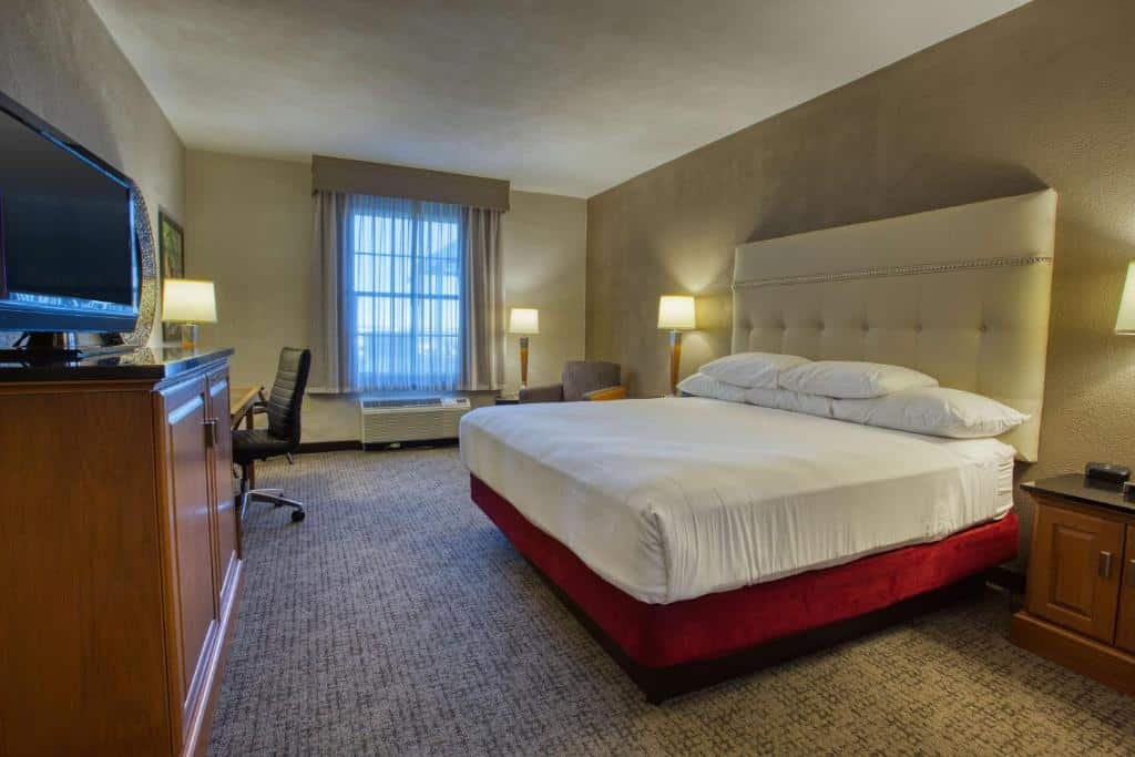 Quarto do Drury Inn & Suites San Antonio Near La Cantera com uma janela com persianas, uma poltrona, uma cama de casal, uma mesa de escritório com uma cadeira giratória, uma móvel com uma televisão em cima