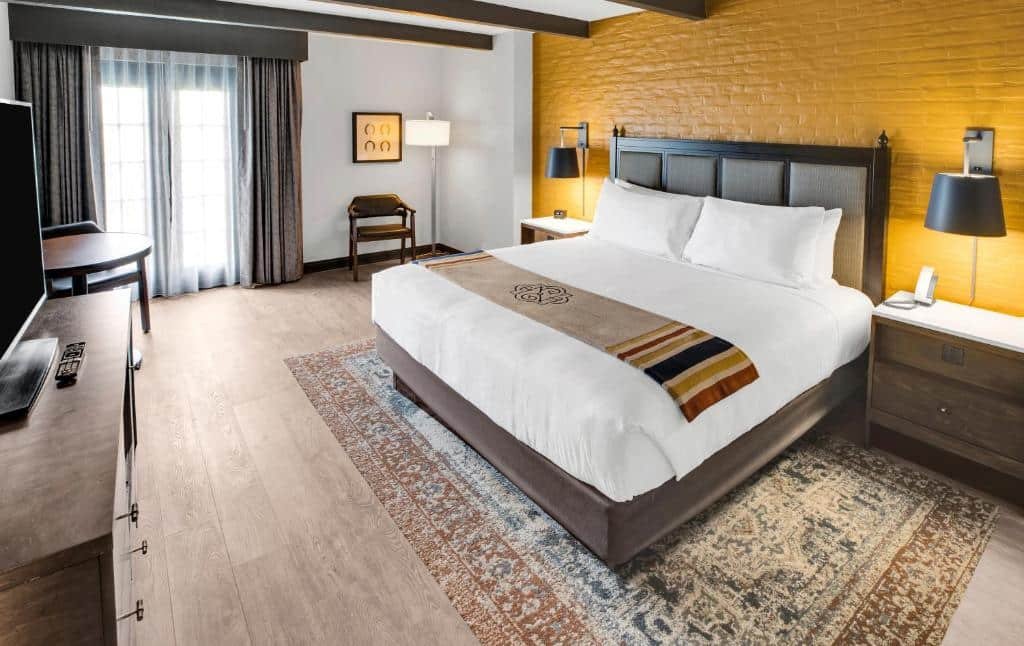 Quarto do Estancia del Norte San Antonio, A Tapestry Hotel by Hilton  com uma cama de casal ampla, uma janela com persiana, uma mesa redonda pequena com duas cadeiras, um móvel com gavetas com uma televisão em cima, e duas mesinhas de cabeceira nas laterais da cama com uma luminária