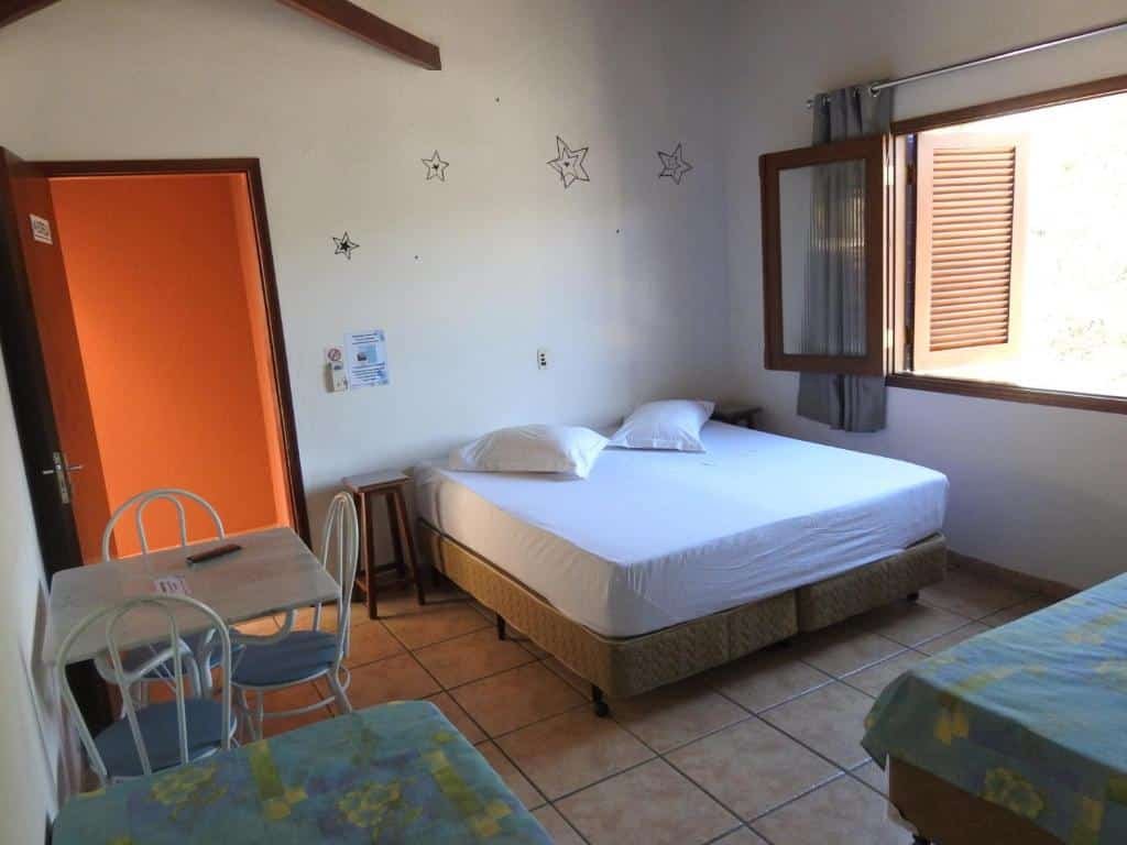 Foto do quarto Estrela da Manhã Pousada, para ilustrar o post sobre pousadas em Águas de São Pedro. Podemos ver uma box cama de casal, do seu lado esquerdo há uma porta e do direito, uma janela. No outro lado do quarto, temos a visão parcial de uma outra cama menor. Uma mesa de mármore com três cadeiras está encostada perto da porta.
