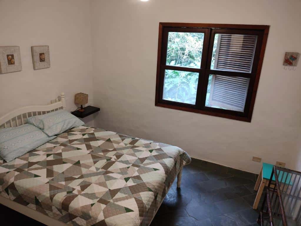 Vista do quarto da Casa aconchegante em Guaeca com cama de casal, luminária do lado esquerdo com janelas de madeiras. Representa airbnb em São Sebastião.