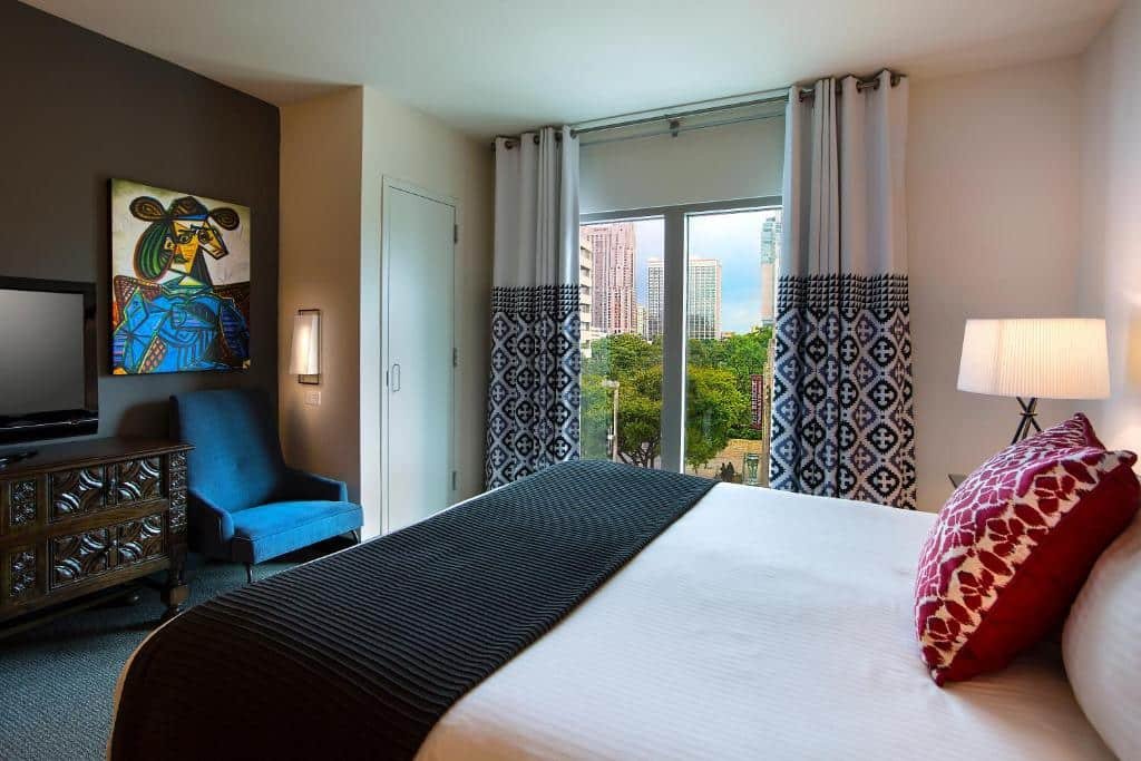 Quarto do Hotel Contessa - Suites on the Riverwalk com uma janela panorâmica com cortinas, uma cama de casal, uma cômoda com uma televisão, e um abajur ao lado da cama, há também um quadro na parede