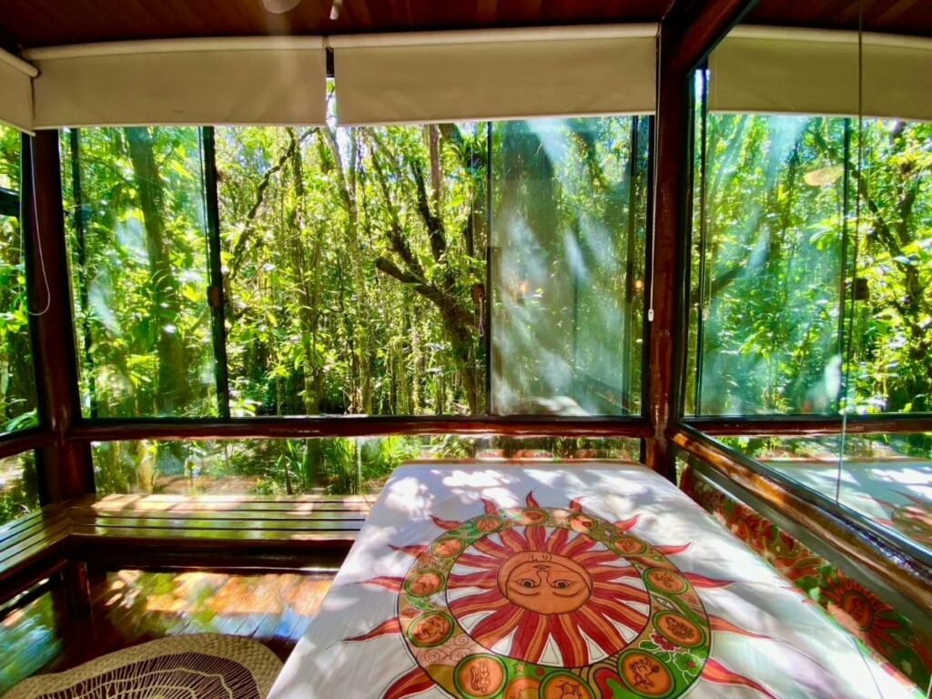 Quarto do Housing 31 durante o dia com cama de casal do lado direito da imagem as paredes são de vidro que tem vista para a natureza.  Representa airbnb em São Sebastião.