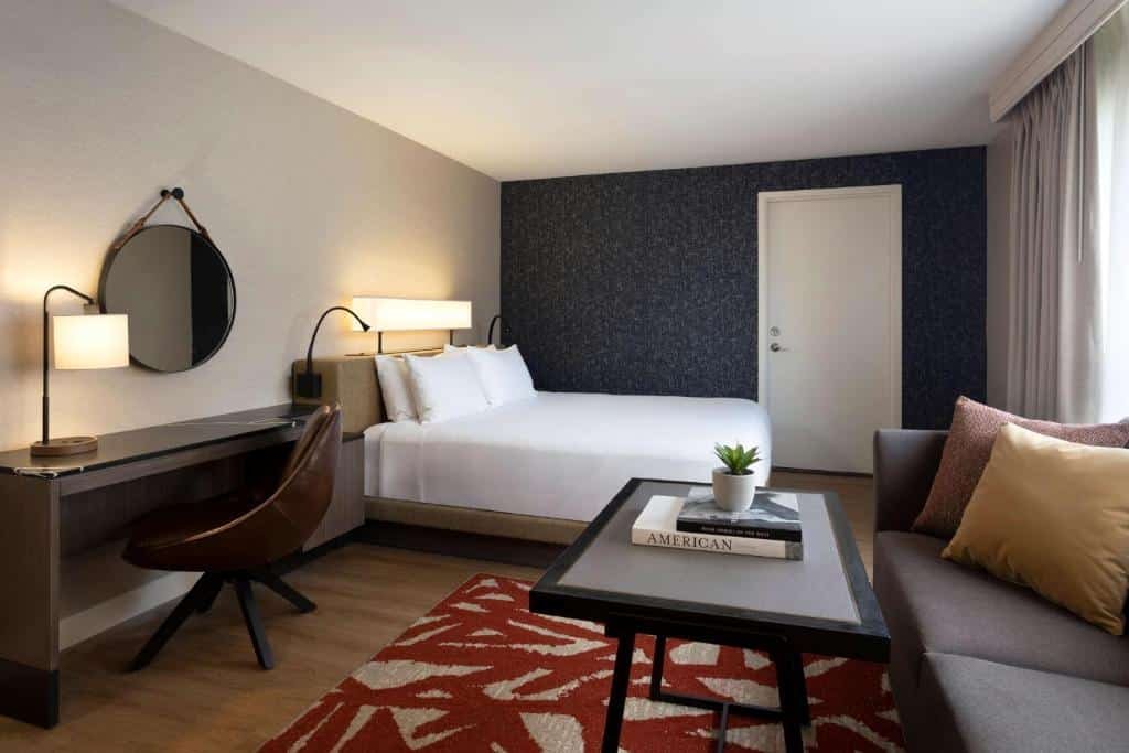 Quarto do Hyatt Regency San Antonio Riverwalk com uma cama de casal, uma mesa de escritório com uma cadeira e um espelho redondo preso na parede acima da mesa, do lado direito, há uma janela, um pequeno sofá e uma mesa de centro