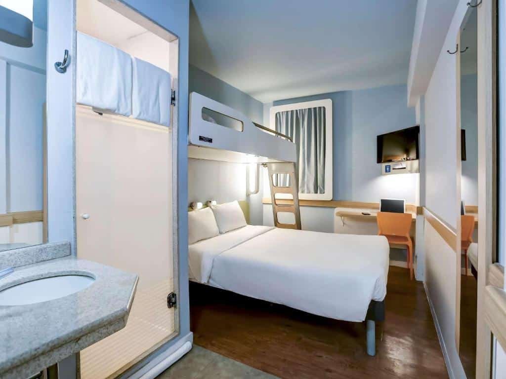Quarto do hotel com uma cama de casal e em cima um beliche de solteiro, uma escrivaninha com cadeira amarela, uma tv e parte da pia do banheiro, ilustrando post hotéis Ibis no Rio de Janeiro.