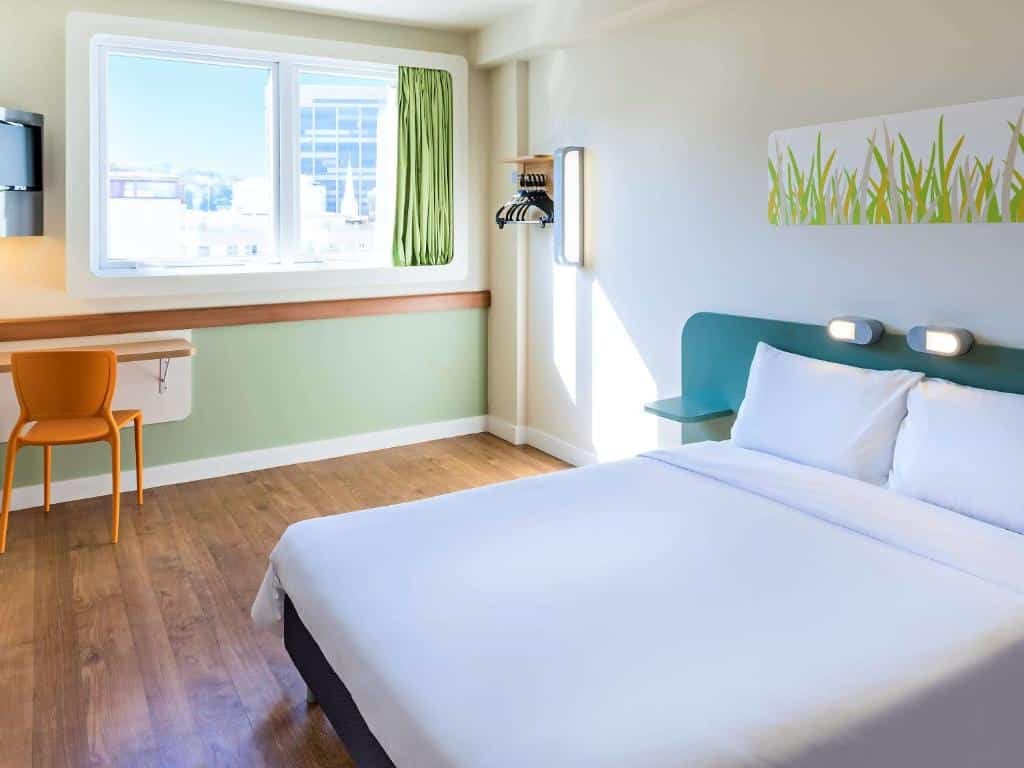 Quarto do hotel com cama de casal branca, chão de madeira, escrivaninha com cadeira amarela, tv, janela de vidro e detalhes de grama verde na parede, ilustrando post hotéis Ibis no Rio de Janeiro.