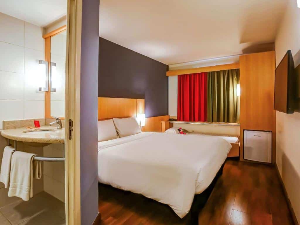 Quarto do hotel com uma cama de casal branca e uma cama de criança, cortina colorida, frigobar, tv e na imagem aparece parte da pia do banheiro, ilustrando post hotéis Ibis no Rio de Janeiro.