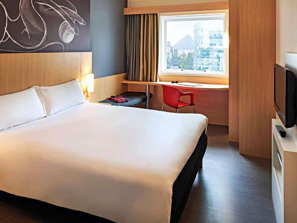 Quarto do hotel com uma cama de casal branca, detalhes brancos na parede preta, escrivaninha com uma cadeira vermelha, janela de vidro e uma tv na parede, ilustrando post hotéis Ibis no Rio de Janeiro.