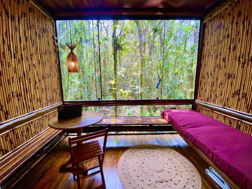 Quarto do Maresias, Chalé H29 em condomínio com cama de solteiro do lado direito da imagem e do lado esquerdo uma mesa redonda com cadeira e notebook ao fundo uma janela panorâmica com vista para o jardim. Representa airbnb em São Sebastião.