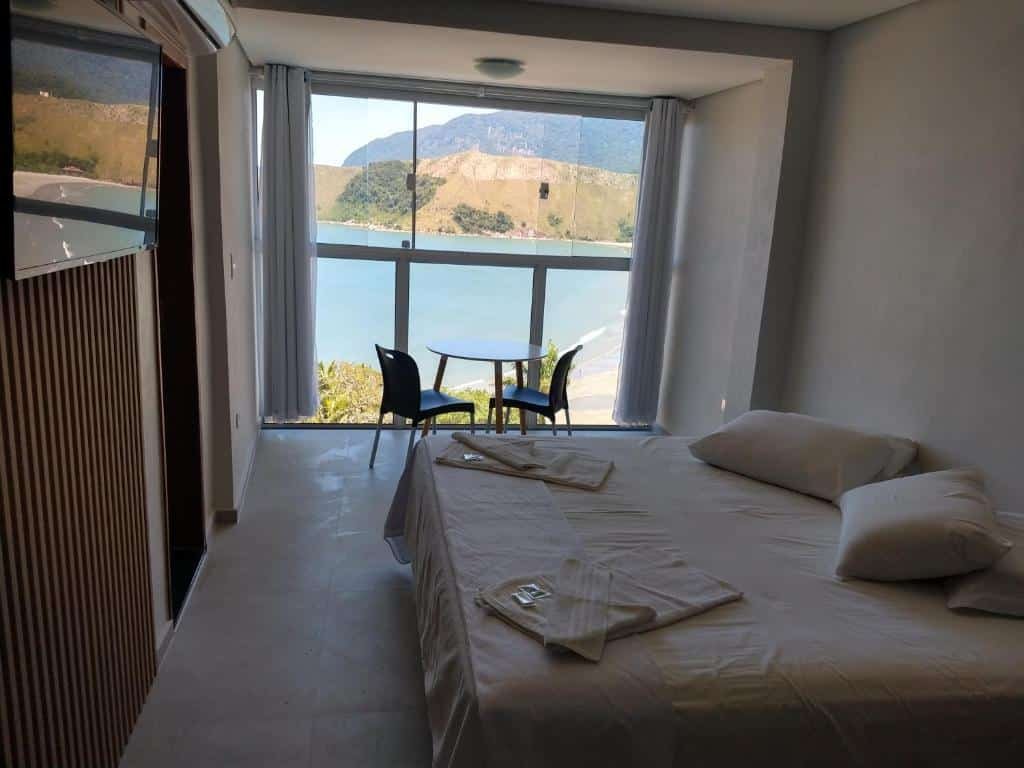 Quarto do Panoramica Residencial com cama de casal do lado direito da imagem, em frente a cama uma parede com TV e do lado direito da cama uma mesa redonda com duas cadeiras com vista para o mar. 