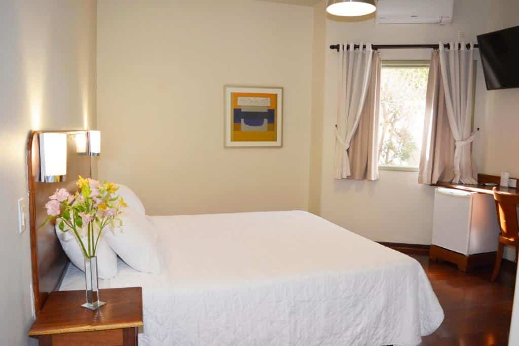 Quarto na Pousada O Garimpo. Uma cama de casal com uma cômoda no canto esquerdo. De frente um frigobar, uma mesa de escritório e uma televisão. Ao lado uma janela.