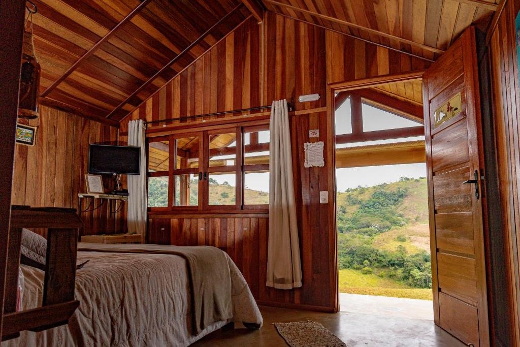 Uma cama de casal no canto esquerdo, com uma televisão na frente, na Pousada Capim Limão. De frente para a cama, a porta da entrada aberta e uma janela, com vista para a serra.