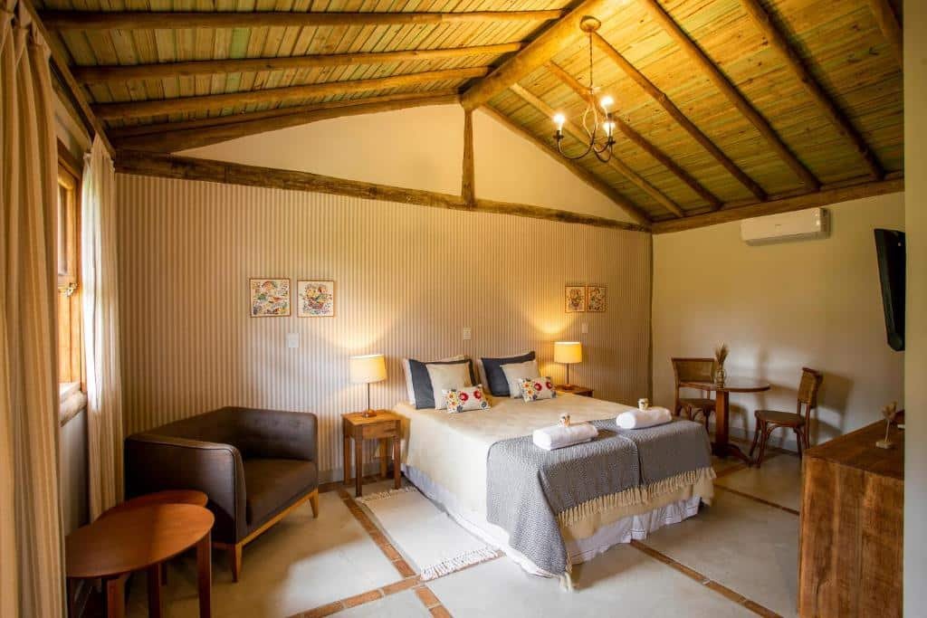 Uma mesa de centro no canto esquerdo, junto com uma poltrona, na Cabanas Portal da Serra. No meio uma cama de casal e no canto direito uma mesa de escritório.