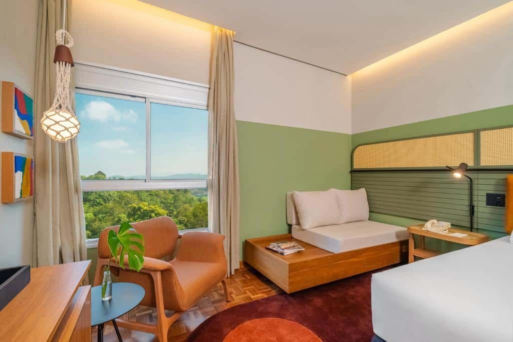 Uma cama no canto direito com uma cômoda e um sofá, no Almenat Embu Das Artes Sao Paulo, Tapestry Collection. No canto esquerdo uma mesa de centro e uma poltrona. Atrás uma janela grande com iluminação natural.