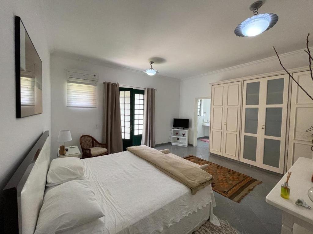 Quarto da pousada Villa Serena Bed & Breakfast. Uma cama de casal no canto esquerdo, um guarda-roupa de frente. Atrás a porta, de um lado uma cadeira e do outro lado uma televisão. Foto para ilustrar post sobre Pousadas em Embu das Artes.