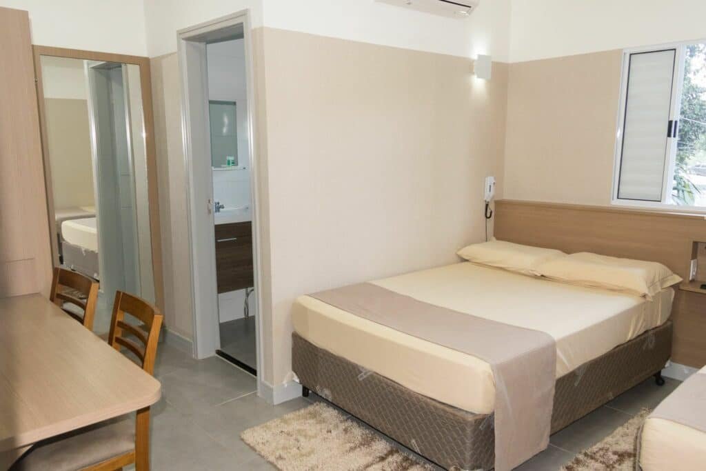 Quarto da Pousada Du Valle. Uma mesa de trabalho com duas cadeiras no canto esquerdo, uma cama de casal e uma cama de solteiro no canto direito. Na frente um espelho e a porta do banheiro. Foto para ilustrar post sobre Pousadas em Embu das Artes.