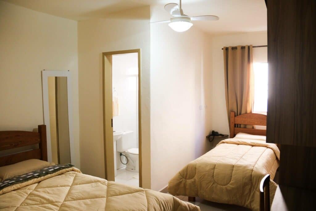 Quarto no Hotel Vale das Artes. Uma cama de casal no canto esquerdo, a porta do banheiro, e uma cama de solteiro no canto direito. Na ponta da foto no lado direito, o armário e a mesa de escritório. Foto para ilustrar post sobre Pousadas em Embu das Artes.