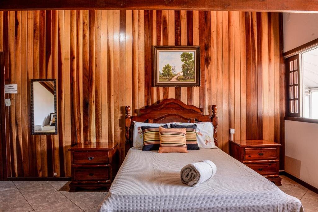 Quarto em pousada da Tina, tudo trabalhado em madeira, uma cama de casal, duas pequenas cômodas, um quadro, uma janela e um espelho, para representar pousadas no Centro de Ubatuba