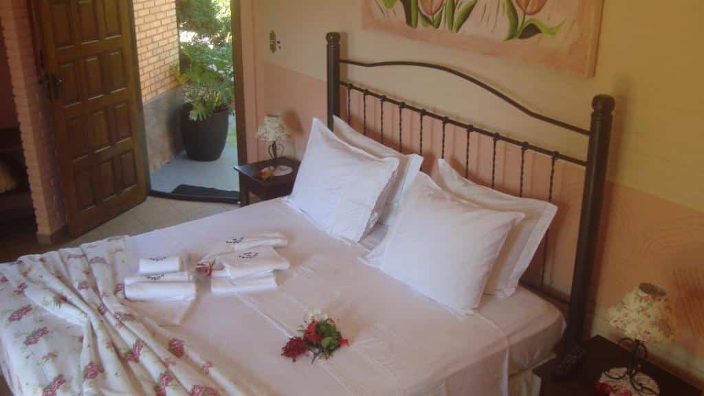 Imagem do quarto na Pousada da Lua, em São Pedro. A cama é de casal e em cima há toalhas de banho e rosas. Em seus lados há uma mesa de cabeceira com abajur em cima. A porta de entrada ao quarto está do lado esquerdo da cama. Representa o post de pousadas em Águas de São Pedro.