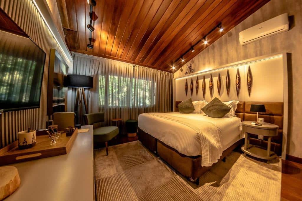 Quarto da Pousada Figueira da Serra com uma cama de casal, chão de madeira com m tapete, móvel de madeira com gavetas e uma televisão, há também uma poltrona e um abajur de chão, além de alguns itens de decoração de madeira